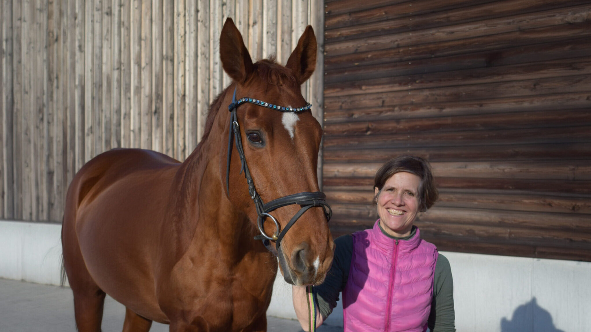 Eine Frau in Reitkleidung lächelt in die Kamera. An den Zügeln hält sie ein braunes Pferd. Im Hintergrund ist eine Reithalle.