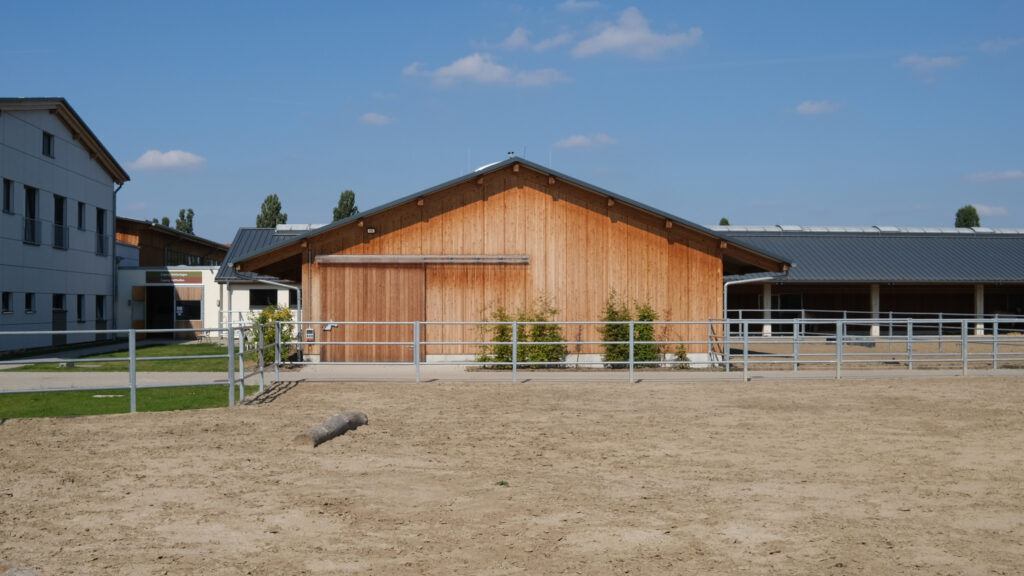 Reitanlage mit Holzstallgebäude, modernem Verwaltungsgebäude und umzäuntem Sandreitplatz bei sonnigem, blauem Himmel.