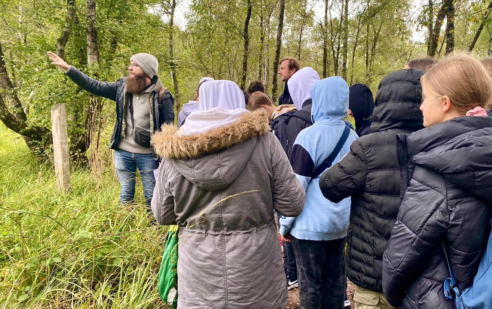 Futurestories Eine Gruppe von Menschen in Jacken steht in einem Wald und hört einem bärtigen Mann mit Hut zu, der gestikuliert und über Moorerhalt und Naturschutz spricht, umgeben von Bäumen und üppigem Grün.